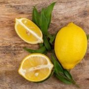 Shows some fresh Tarragon leaves on a wooden surface with a whole lemon on the right side of it and pieces of a cut lemon on the left.